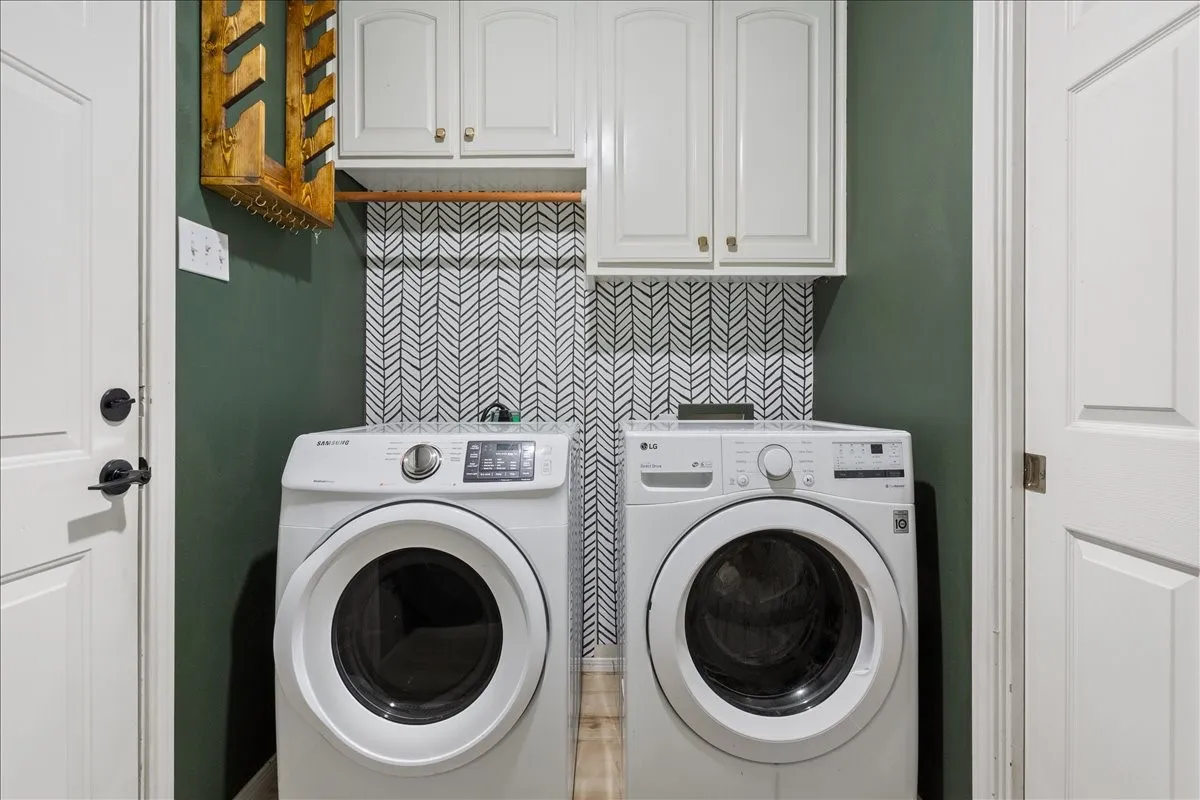 Dedicated laundry area featuring a patterned backsplash, overhead cabinetry with brass-finish hardware, a wooden hanging rod, and contrasting accent wall