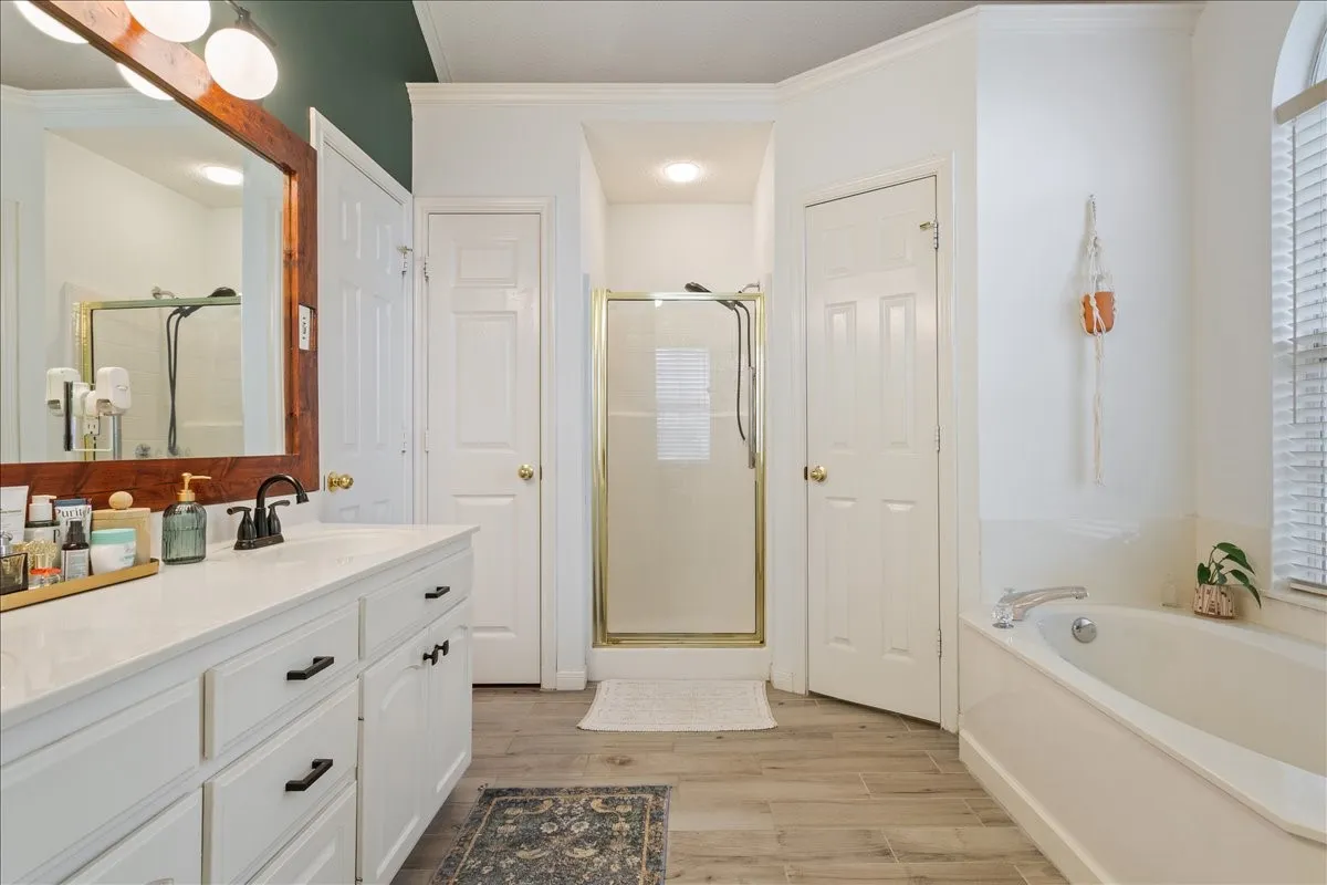 Bathroom featuring a white vanity with black hardware, a wood-framed mirror, a built-in bathtub, a glass-enclosed shower, and wood-finish flooring