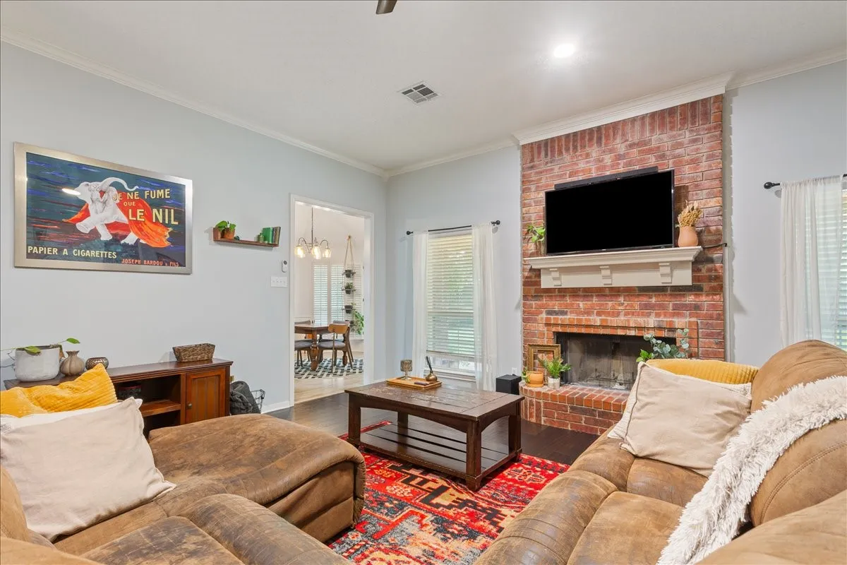 Living area featuring a brick fireplace with a white mantel, dark wood-finish flooring, light-colored walls, crown molding, and a visible doorway to an adjacent room