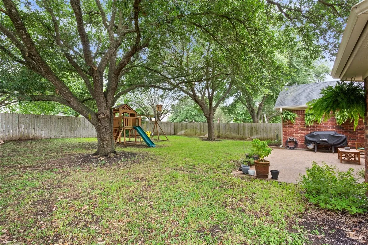 Expansive backyard featuring mature trees, a wooden privacy fence, a brick exterior with a dark shingle roof, and a concrete patio