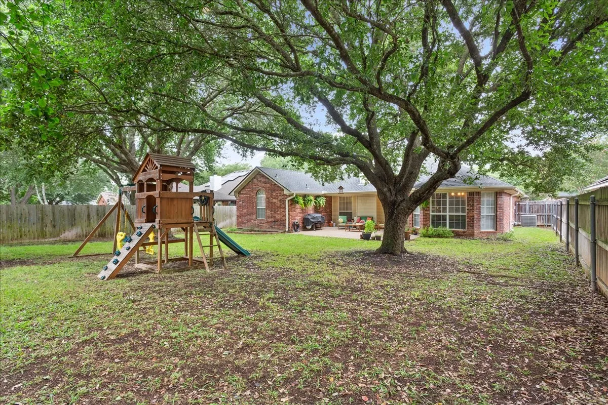 Expansive fenced backyard featuring a large mature tree providing natural shade, a brick exterior, and a concrete patio
