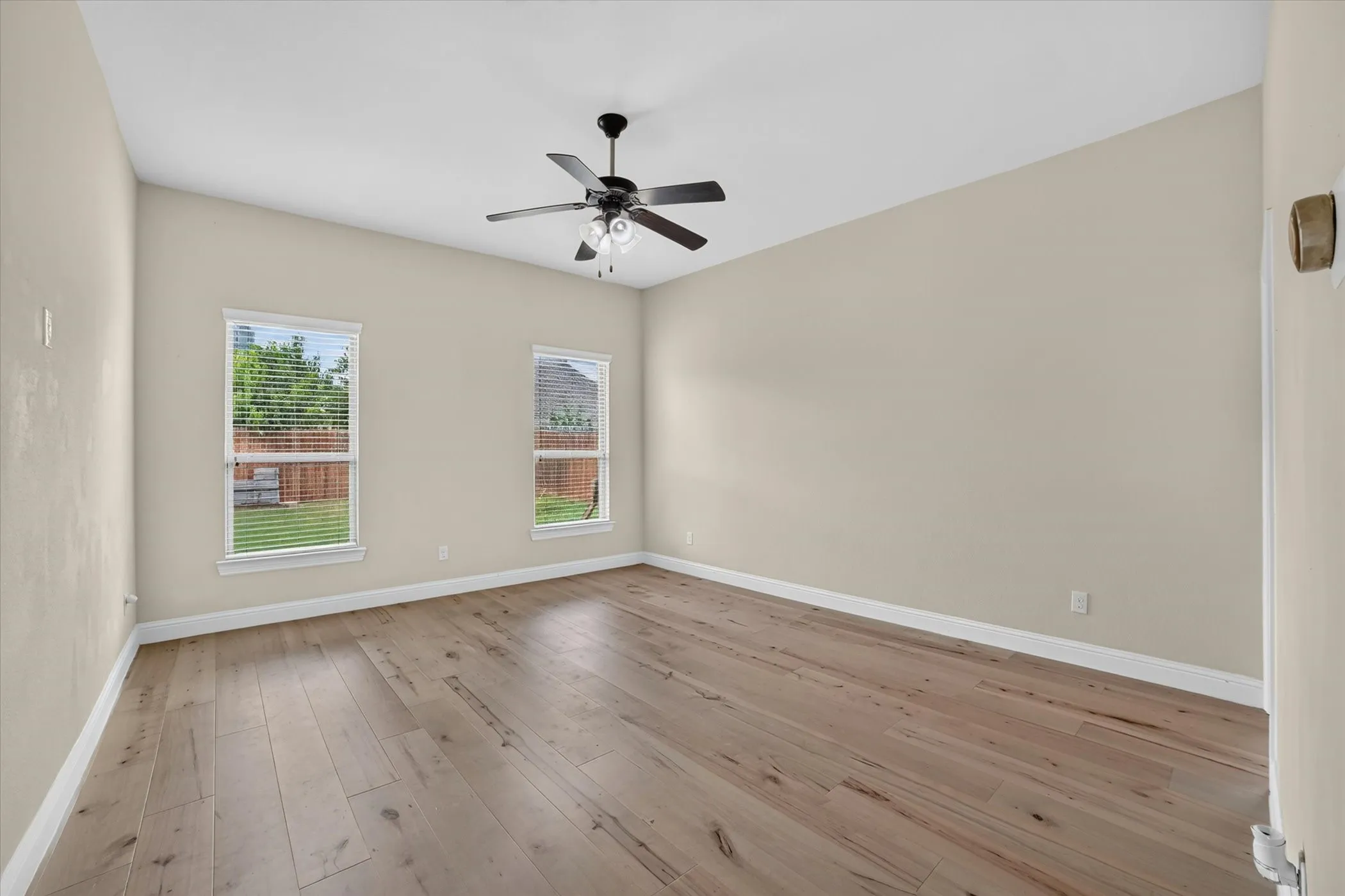 Spacious room featuring wood-finish flooring, two windows with blinds, a ceiling fan with integrated lighting, and neutral wall tones