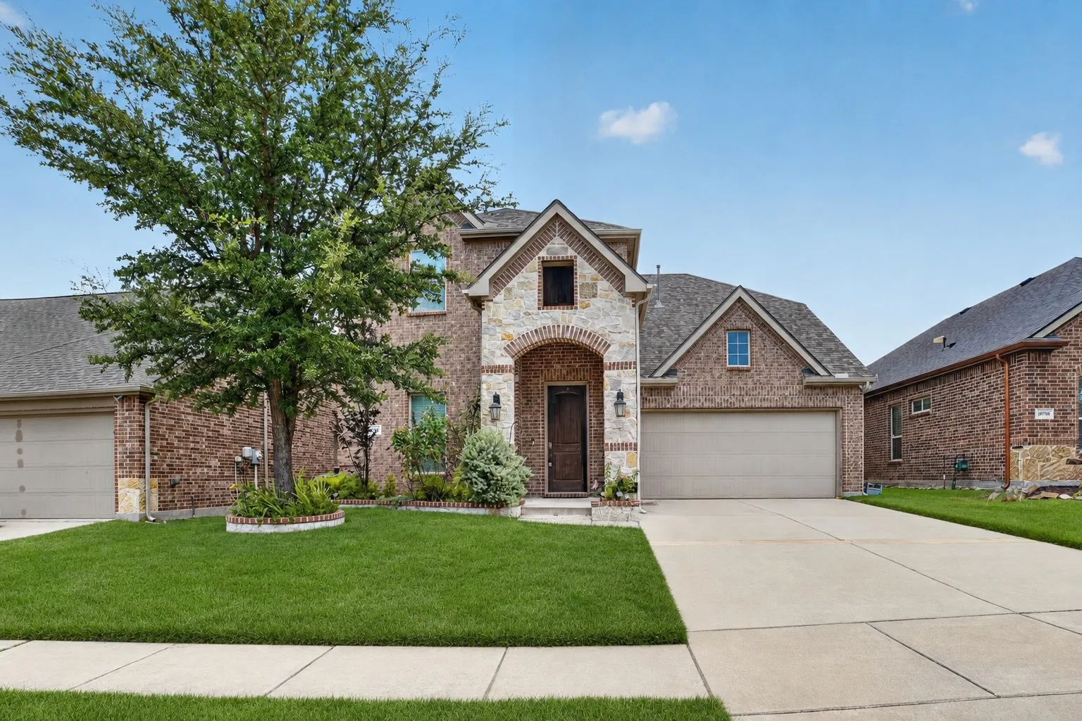Brick and stone exterior featuring an arched entry, mature tree, manicured lawn, and an attached two-car garage