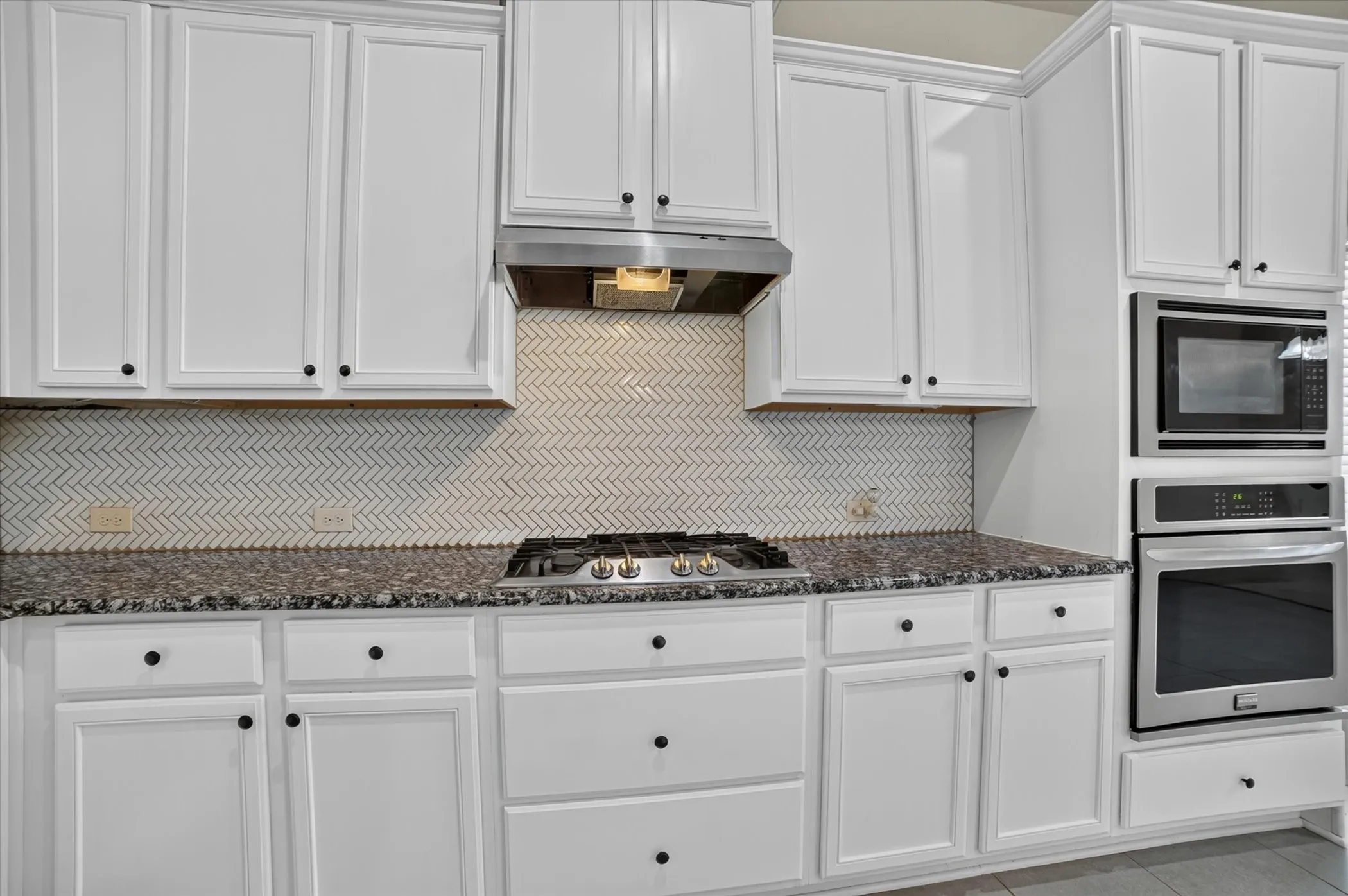Kitchen with white shaker cabinetry, dark countertops, a herringbone tile backsplash, and stainless steel appliances including a gas range and built-in oven and microwave