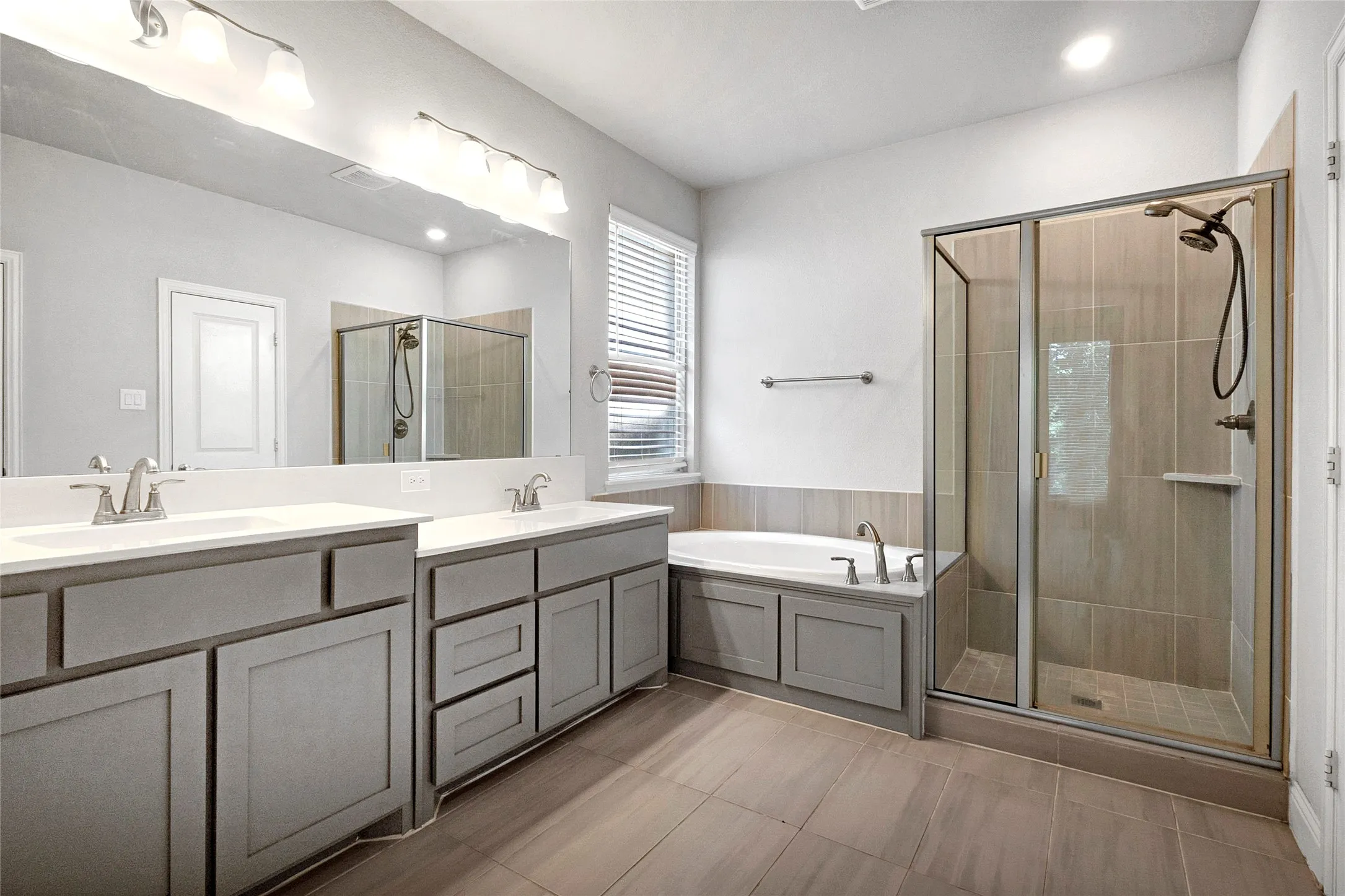Spacious bathroom featuring a dual vanity with light gray cabinetry, a walk-in shower with glass enclosure, a built-in soaking tub, and tile flooring