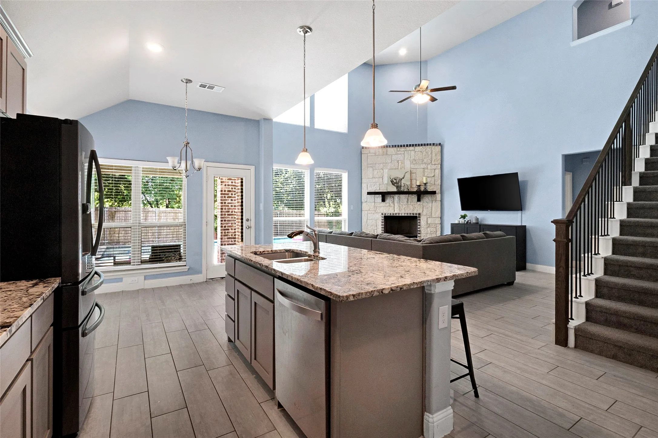 Open-concept living space featuring a central kitchen island with granite countertops, wood-finish flooring, a stone-faced fireplace, and a staircase with wrought iron balusters