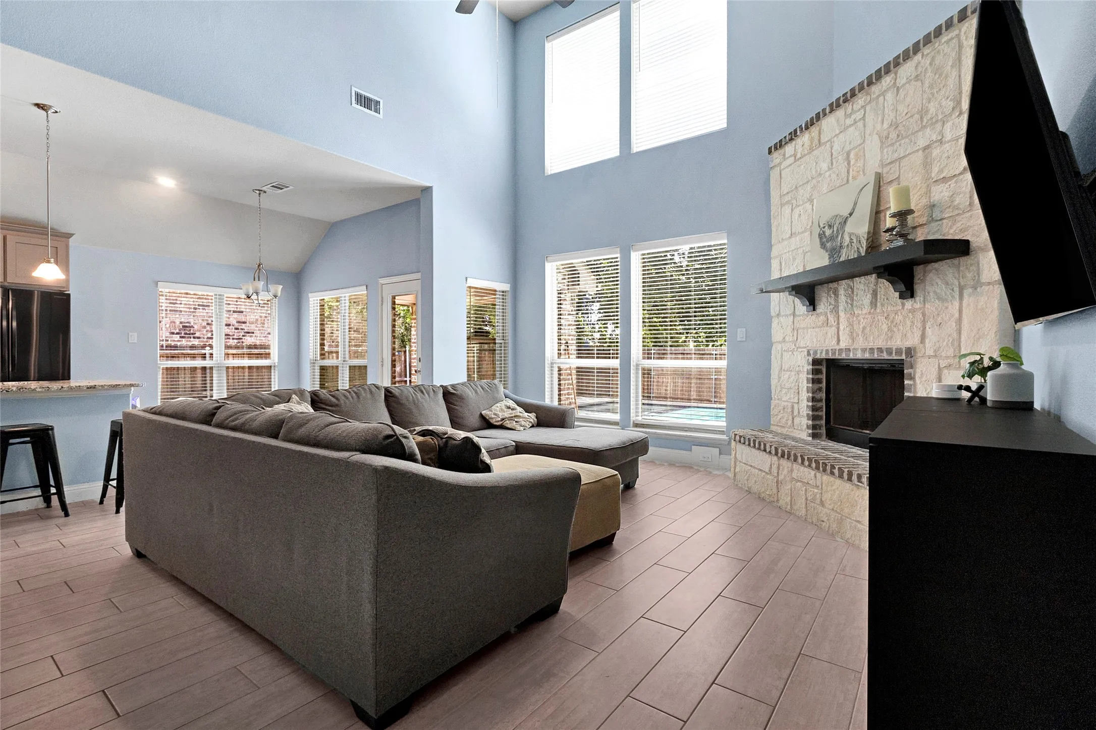 Two-story living area with high ceilings featuring wood-finish flooring, a stone-faced fireplace with a dark wood mantel, and multiple windows providing outdoor access