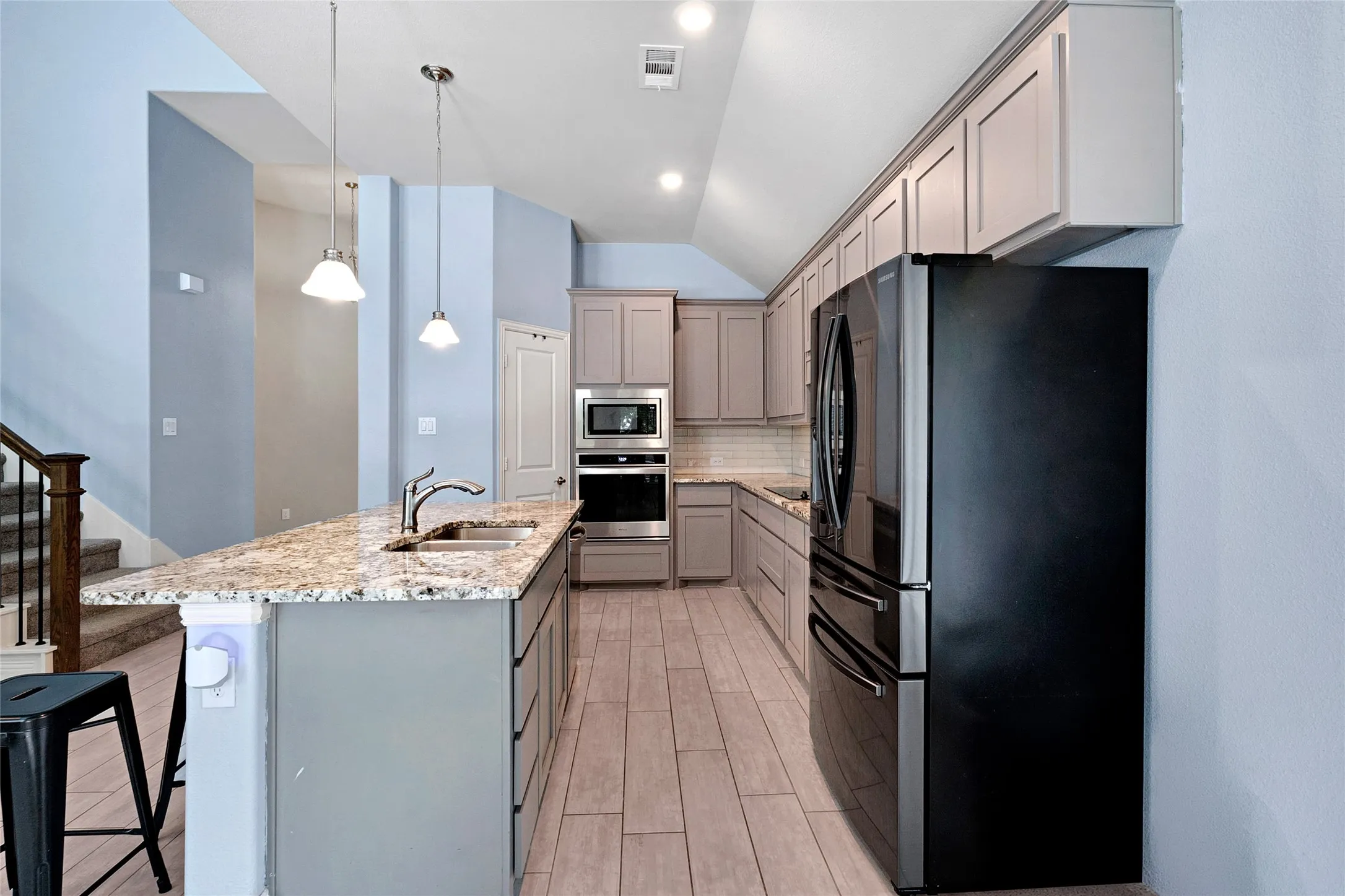 Modern kitchen featuring a large island with a double basin sink, light-toned granite-style countertops, shaker-style cabinetry, stainless steel appliances, and wood-finish tile flooring