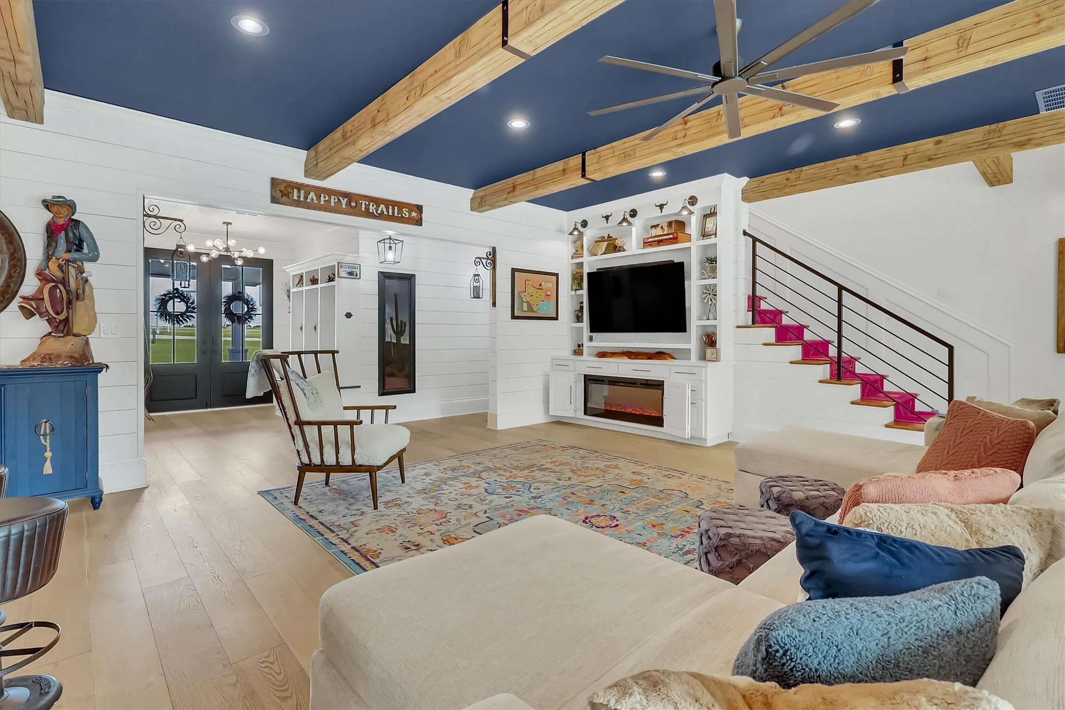 Living area featuring a blue ceiling with exposed wood beams, wood-finish flooring, a built-in media center with an electric fireplace, shiplap wall accents, and a staircase with a black metal railing