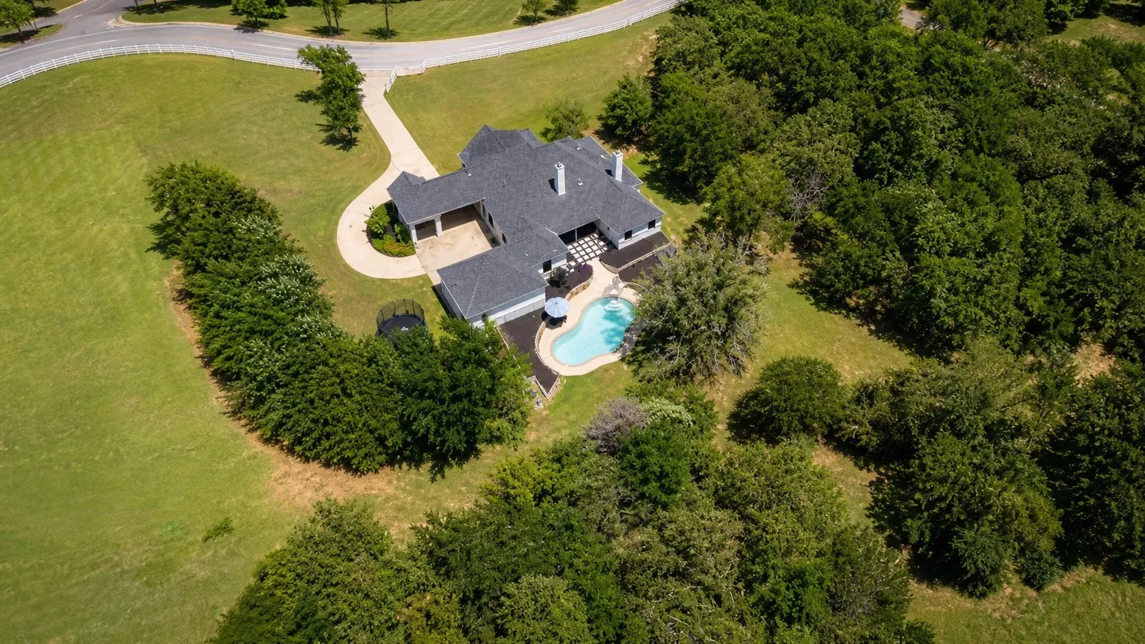 Expansive property featuring a residence with a dark shingle roof, a curving driveway, an attached two-car garage, a checkered patio, and an outdoor swimming pool