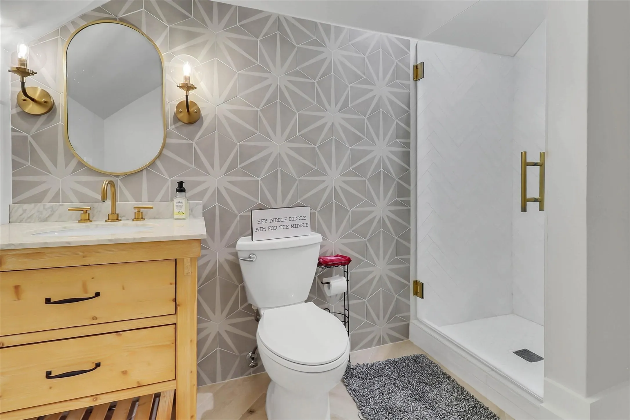 Bathroom featuring a geometric tiled accent wall, glass-enclosed shower with brass hardware, wood-finish vanity with a stone countertop, and brass wall-mounted sconces