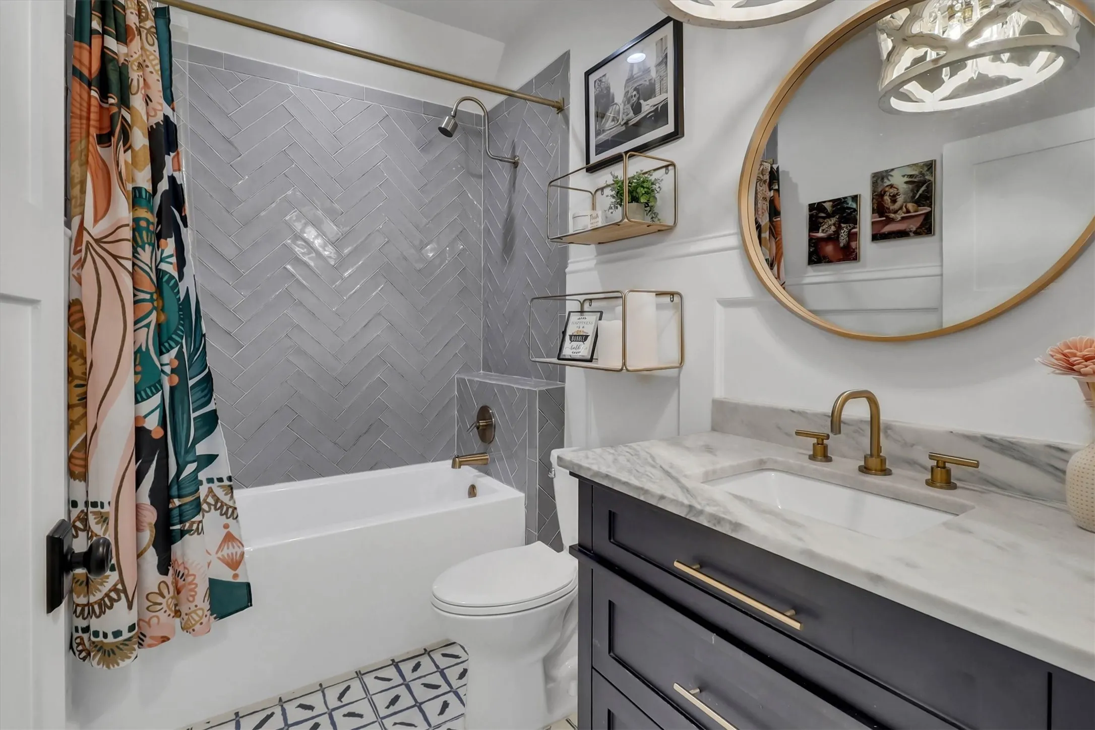 Bathroom featuring a white tub with a herringbone-patterned tile surround, a dark vanity with a light stone countertop, a circular mirror, and a brass-finish faucet