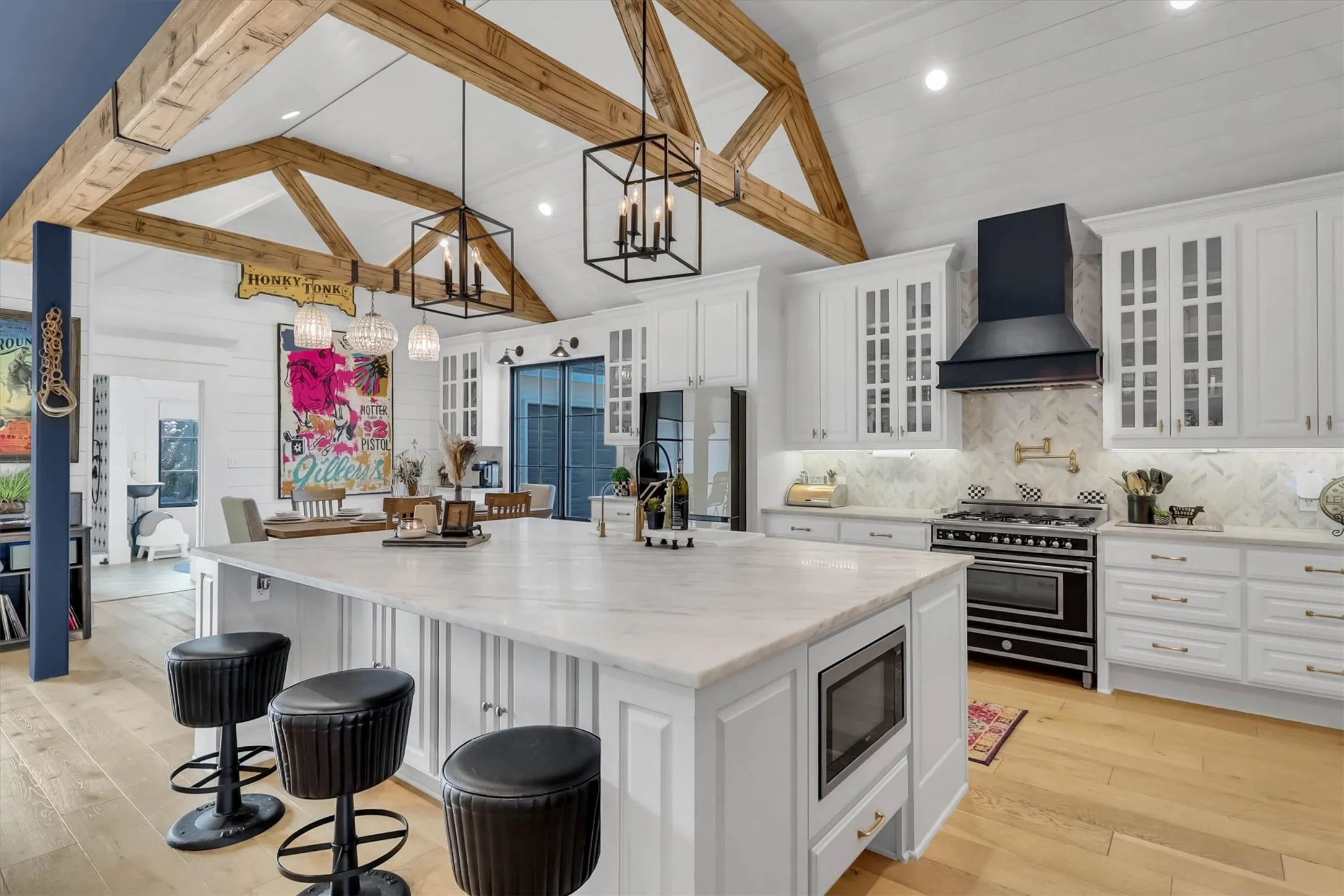 Vaulted ceiling with exposed wood beams, white cabinetry with glass-front uppers, a large kitchen island with integrated microwave, a black range with coordinating hood, and light wood-finish flooring