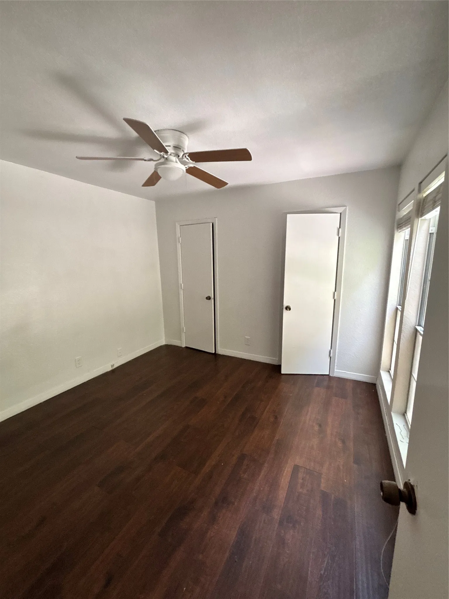 Interior room featuring wood-finish flooring, white walls, two white doors with dark hardware, and a ceiling fan with light