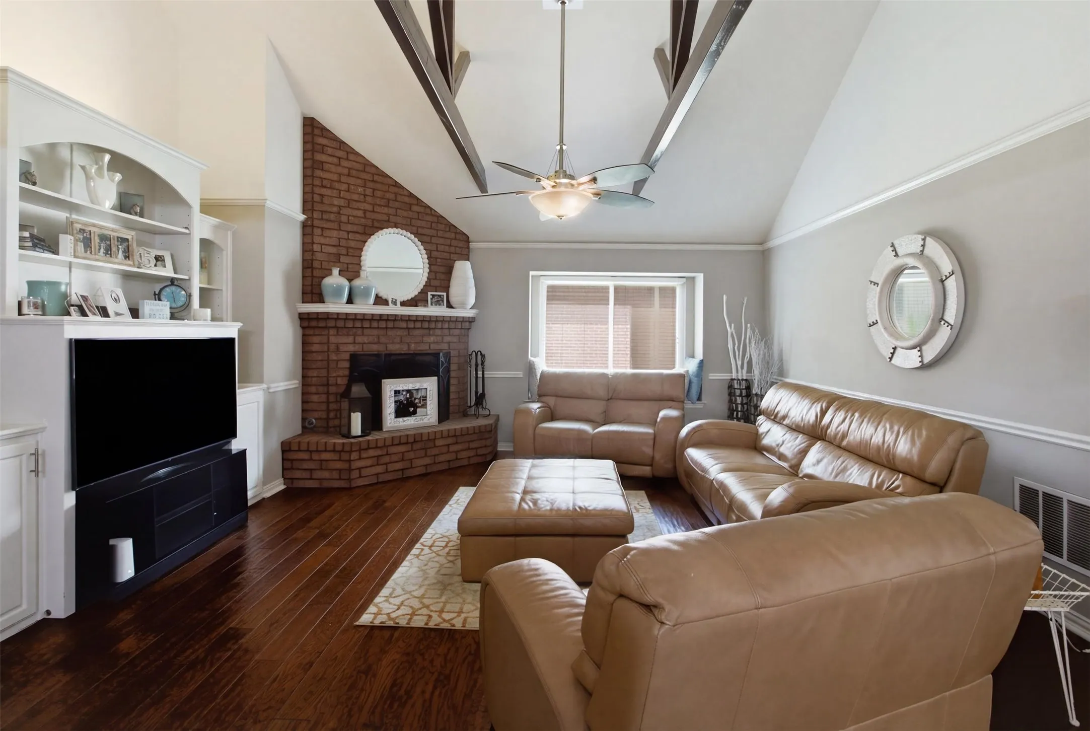 Living area featuring vaulted ceilings with exposed wood beams, a brick fireplace with a mantel, dark wood-finish flooring, built-in shelving, and a ceiling fan with integrated lighting