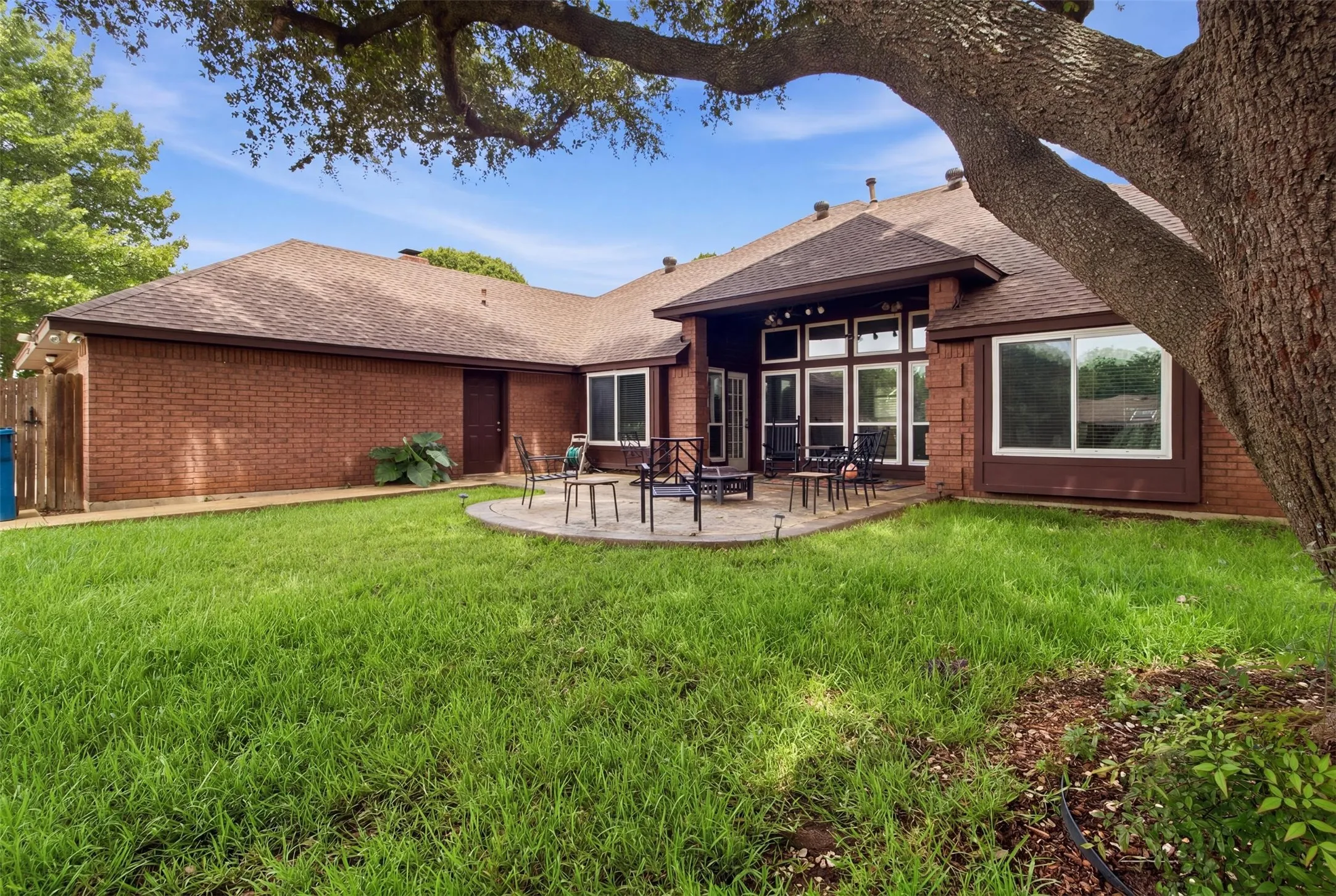 Expansive backyard featuring a manicured lawn, a large circular paver patio, brick exterior, and a prominent mature tree providing natural shade