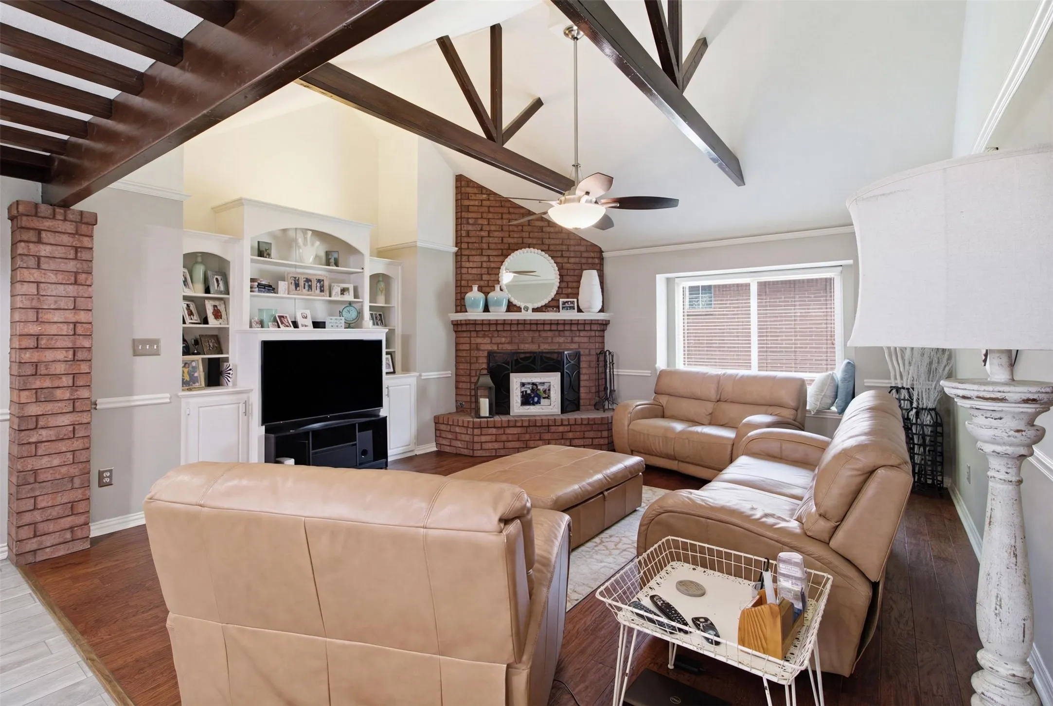Living area featuring vaulted ceilings with exposed wood beams, a brick fireplace, built-in shelving, wood-finish flooring, and a ceiling fan