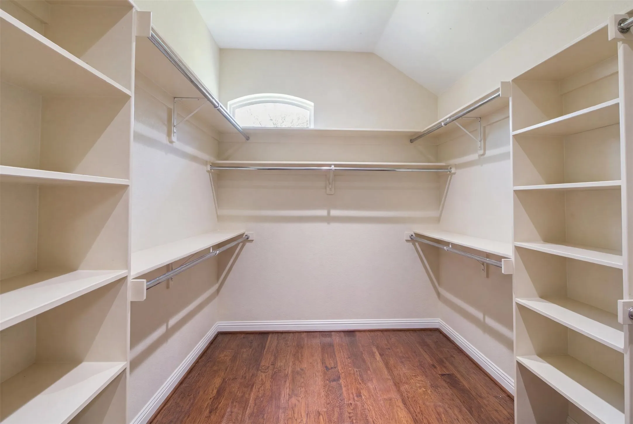 Walk-in closet featuring extensive built-in shelving, multiple hanging rods, rich wood-finish flooring, and a decorative arched window