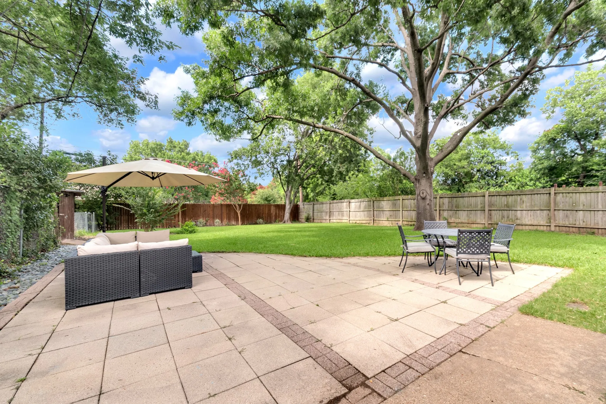 Expansive backyard featuring a large paved patio with brick edging, a generous lawn area, mature trees, and a wood privacy fence