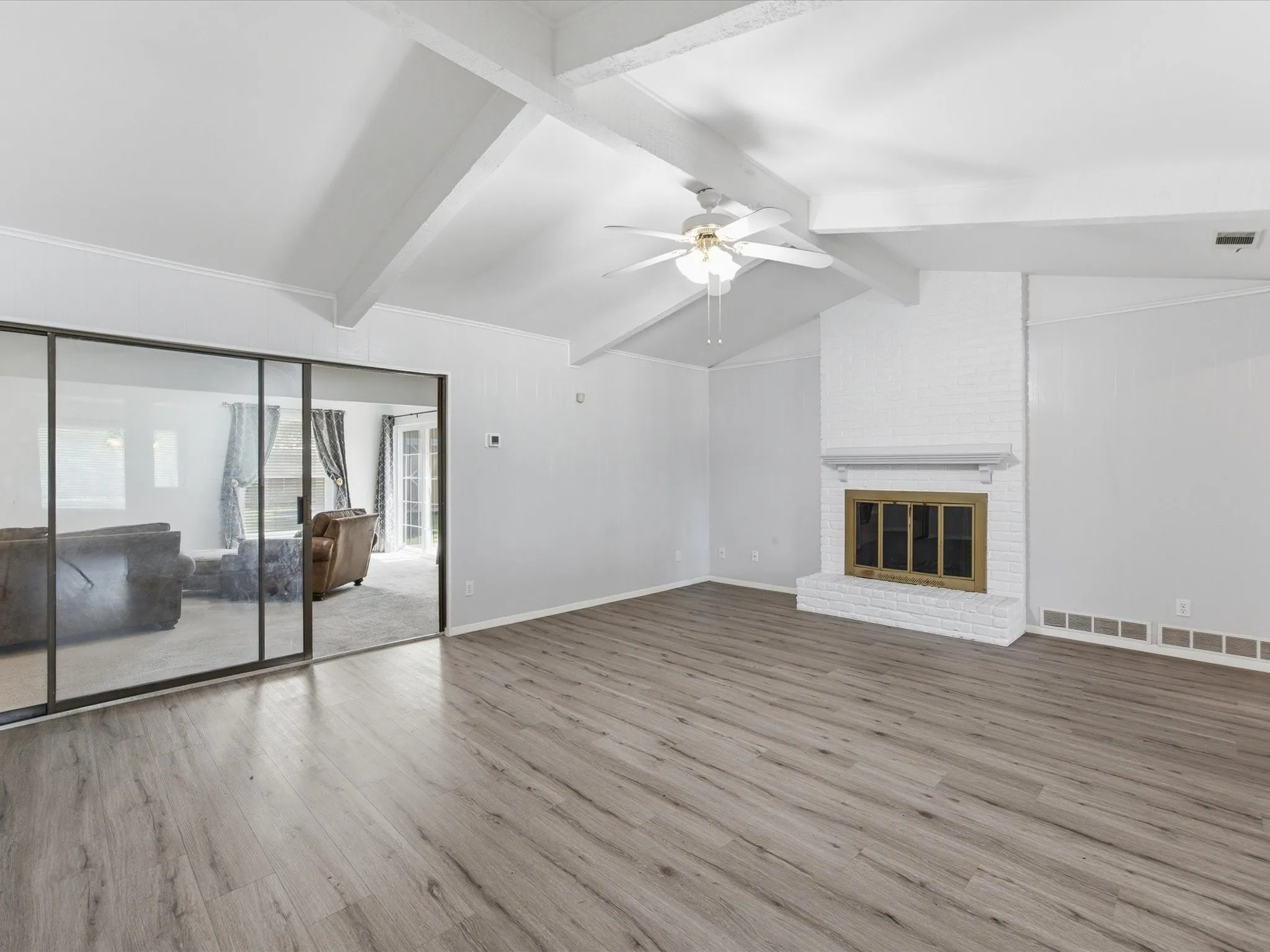 Spacious living area featuring wood-finish flooring, a white brick fireplace with a decorative mantel, and a vaulted ceiling with exposed beams