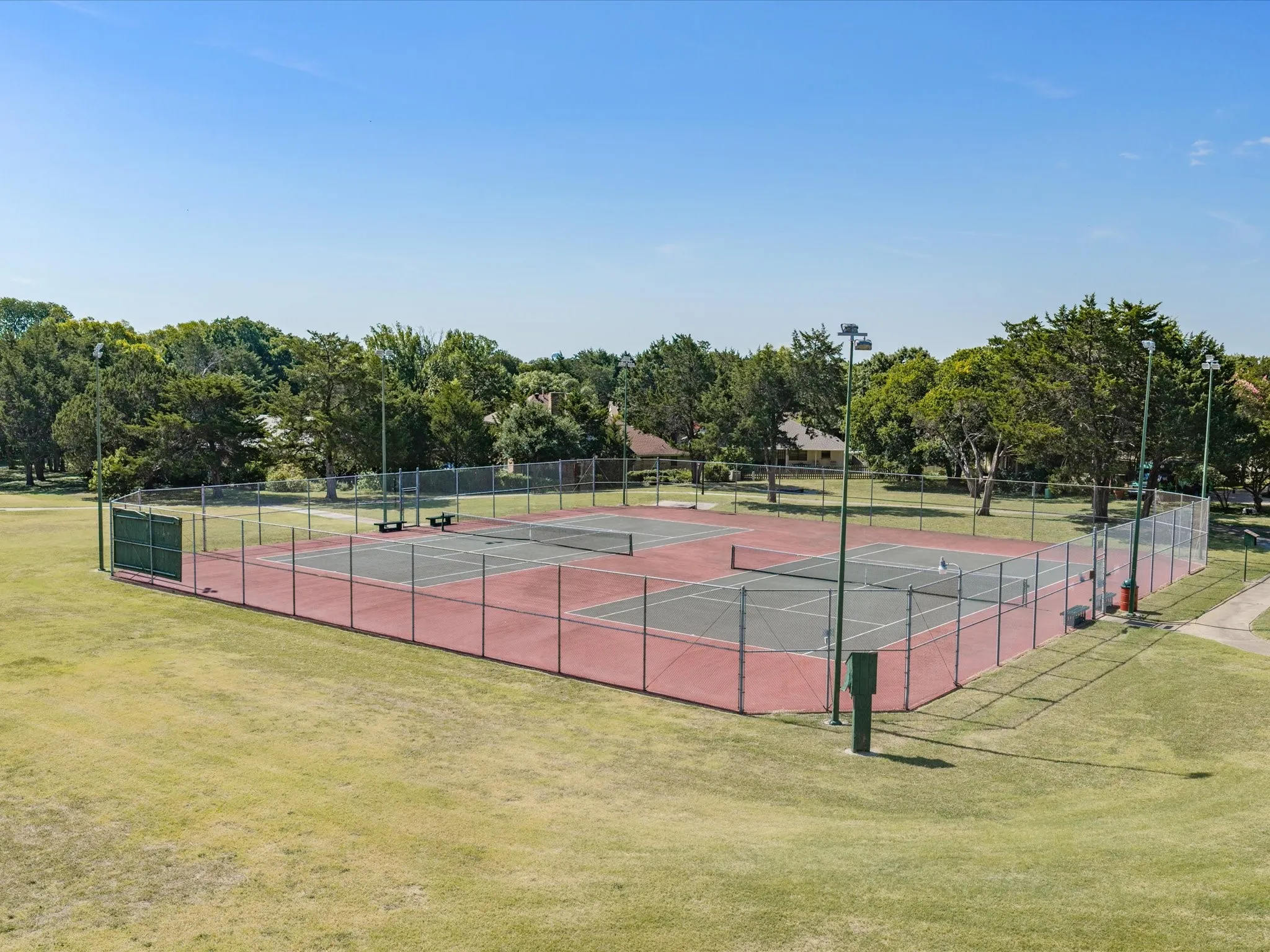 Fenced tennis courts featuring a red and grey surface, surrounded by mature trees and a grassy expanse