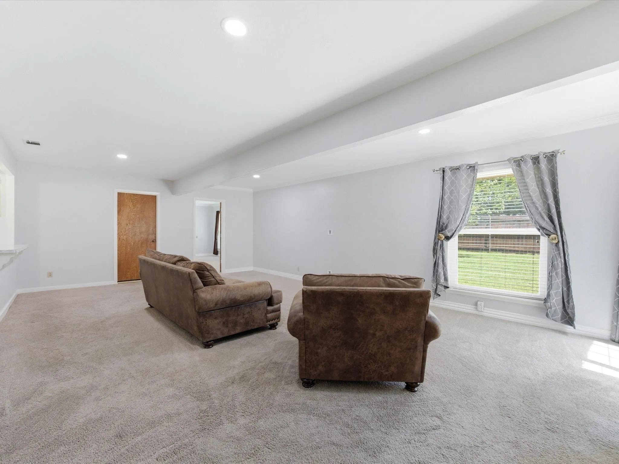 Spacious room featuring light-colored carpeting, recessed lighting, and a large window with blinds overlooking a fenced backyard