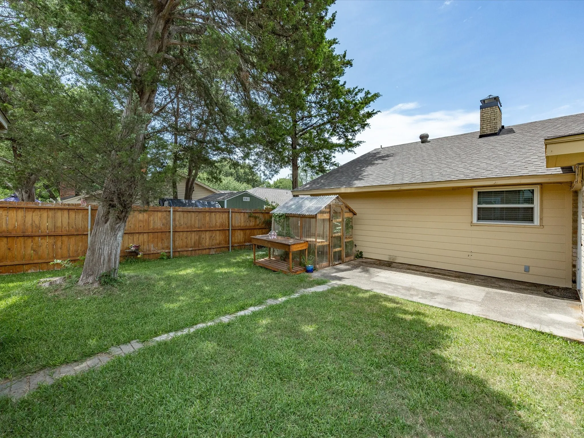 Expansive grassy yard featuring a wood privacy fence, mature trees, a concrete patio, and a compact greenhouse