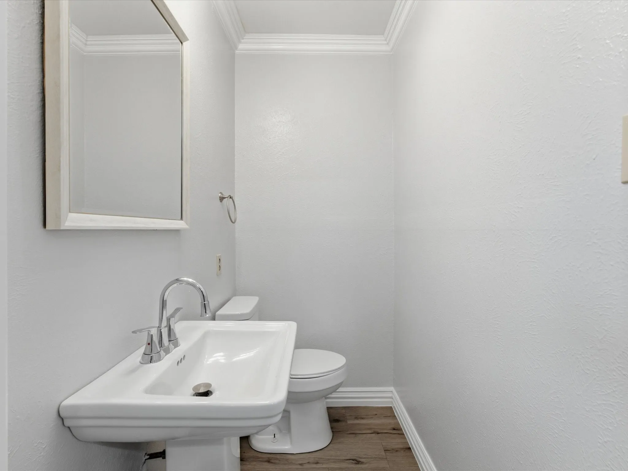 Pedestal sink with chrome fixtures and framed mirror, complemented by wood-finish flooring, white baseboards, and crown molding