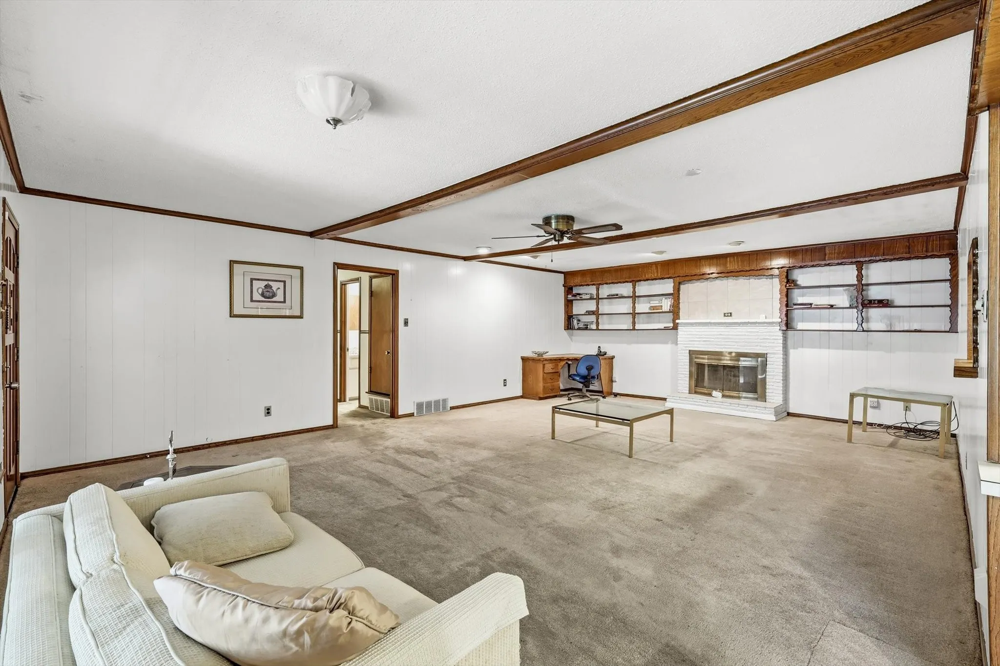 Spacious room featuring a white brick fireplace with a metallic insert, built-in shelving, wood-beam ceiling details, and a ceiling fan