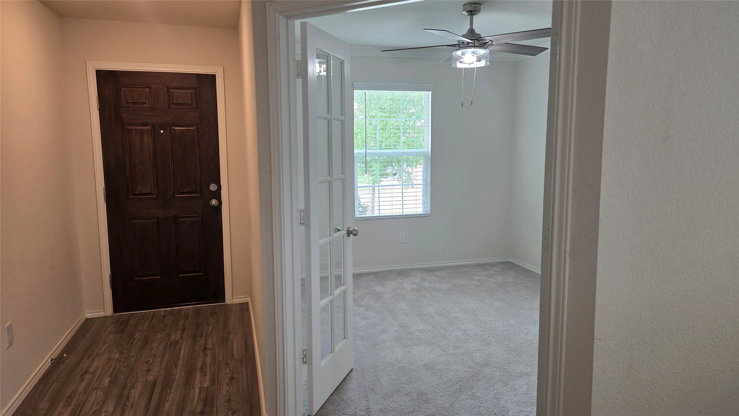 Entryway with wood-finish flooring and a dark paneled door, featuring a room with french doors, carpeted flooring, a window with horizontal blinds, and a ceiling fan with integrated lighting