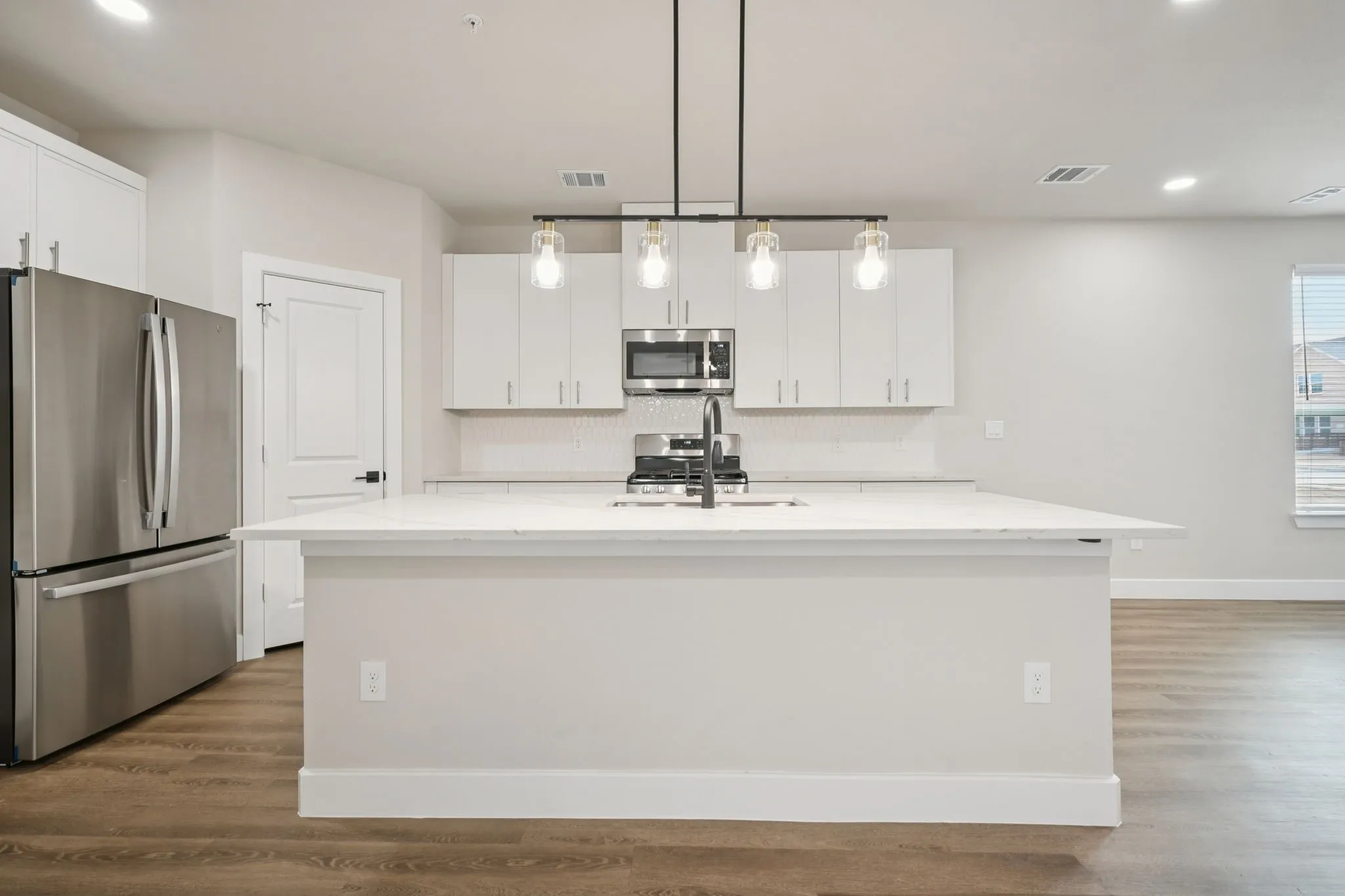 Modern kitchen featuring a central island with a sink, stainless steel appliances, white cabinetry, wood-finish flooring, and pendant lighting