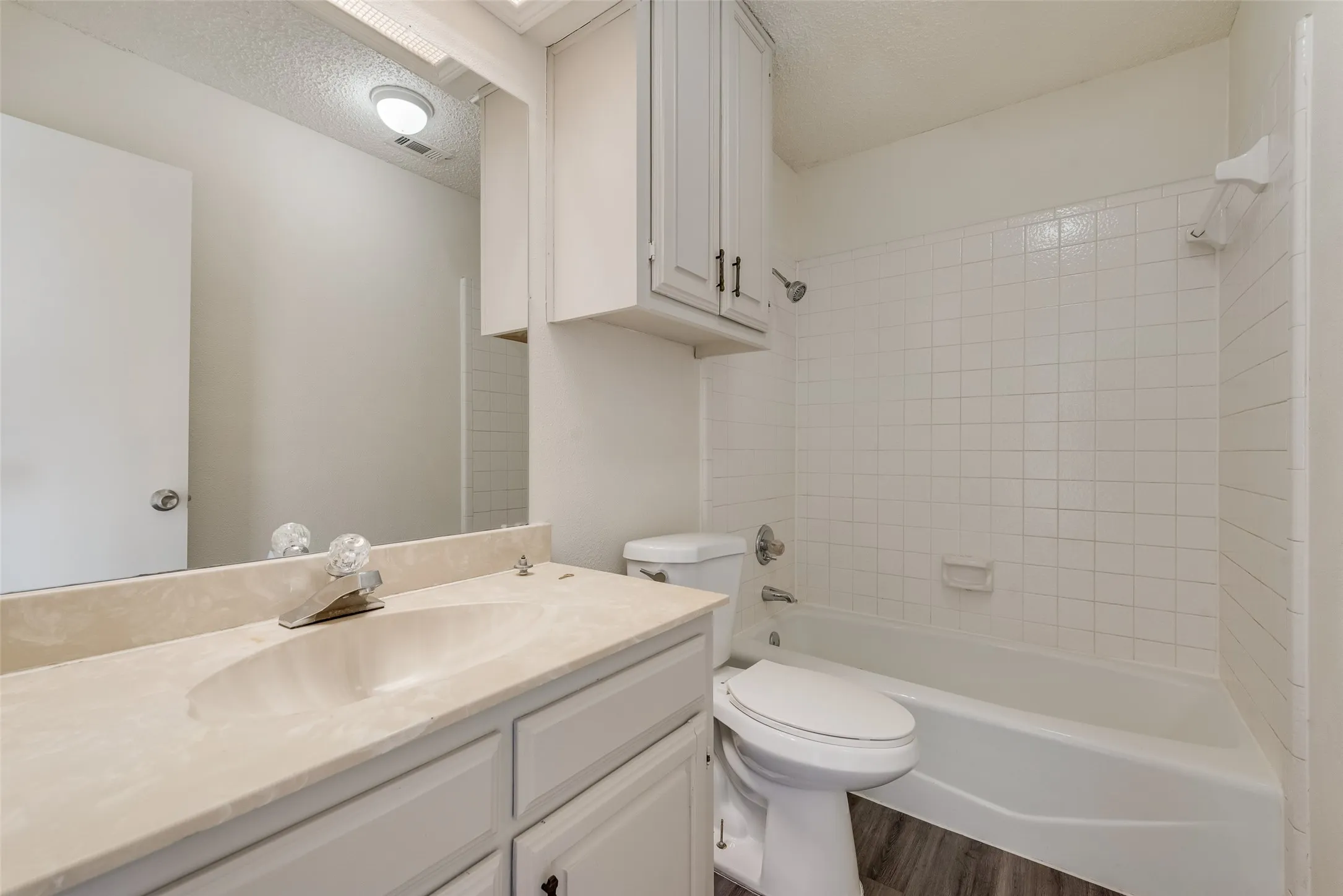 Bathroom featuring a single vanity with an integrated sink, a large mirror, white cabinetry, a built-in tub with white tile surround, and wood-finish flooring