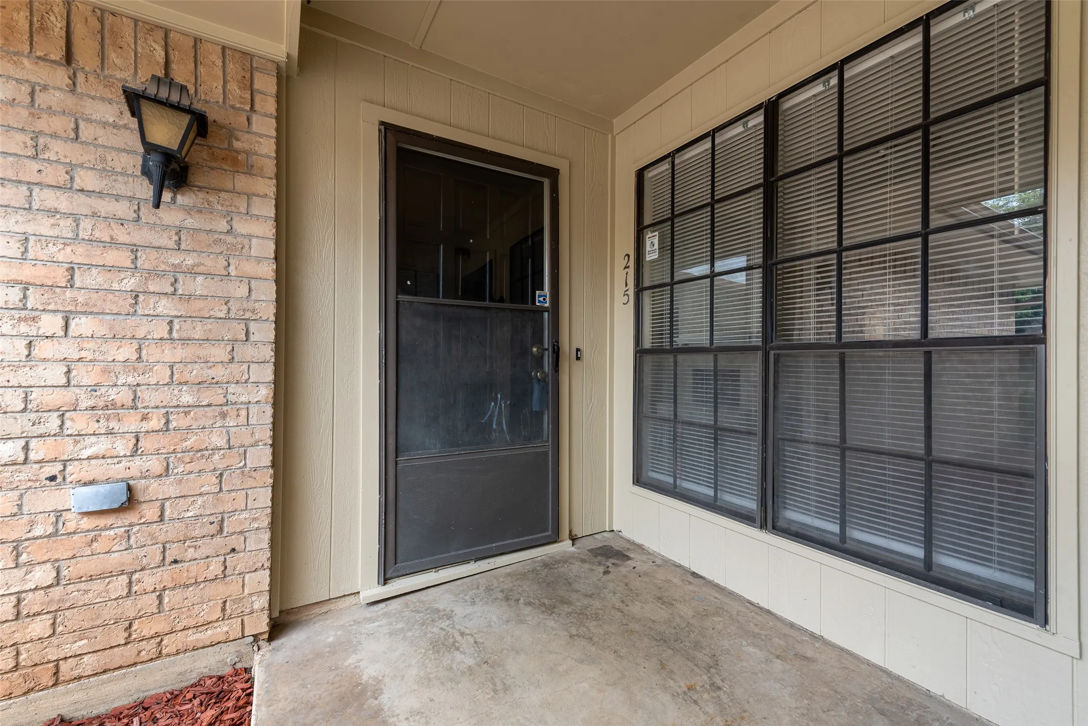 Entryway featuring a security screen door, large windows with horizontal blinds, and a brick exterior wall with an exterior light fixture