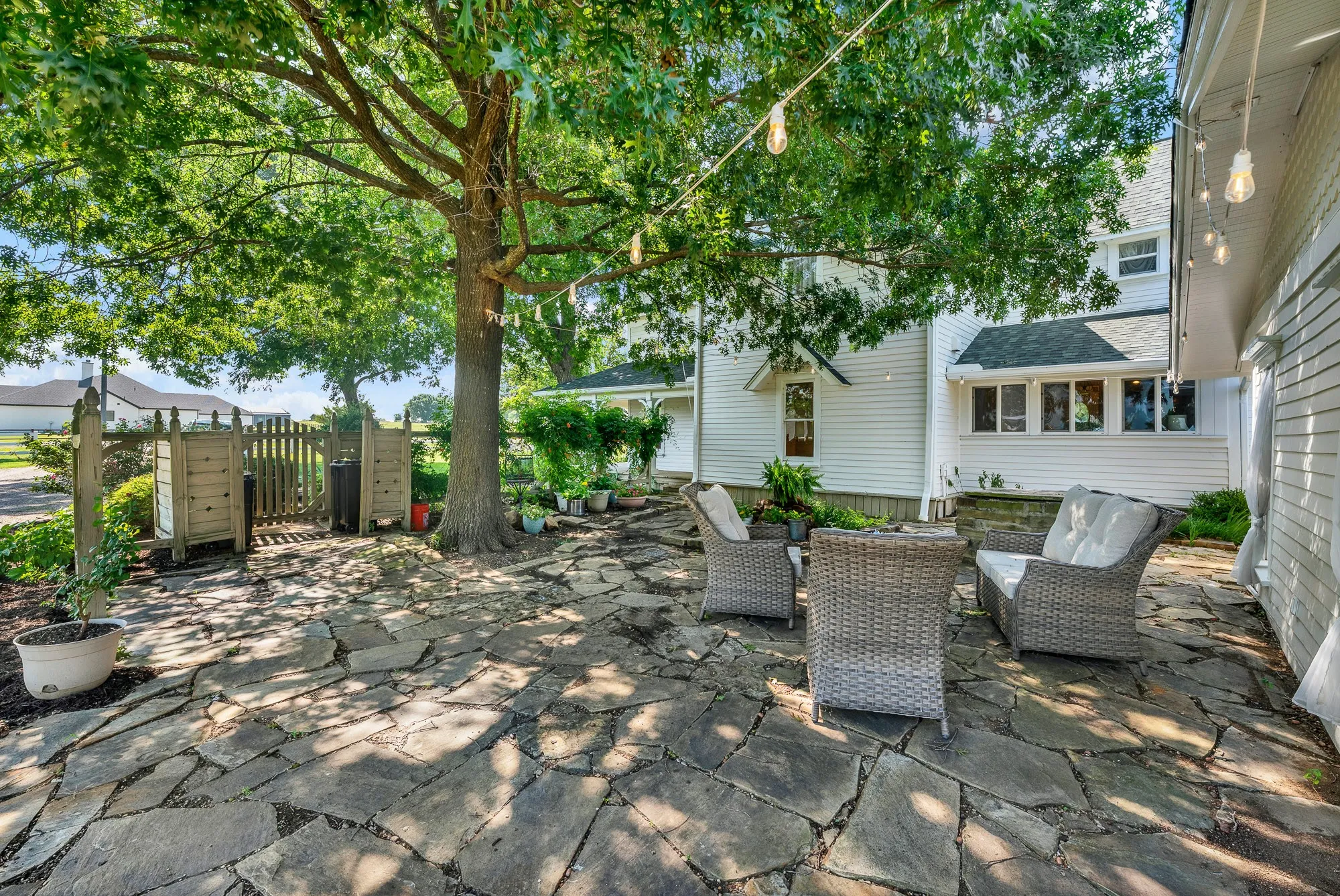 A charming flagstone courtyard creates a peaceful outdoor retreat beneath the shade of mature oak trees. String lighting, lush landscaping, and ample seating provide the perfect setting for morning coffee, quiet evenings, or entertaining guests.