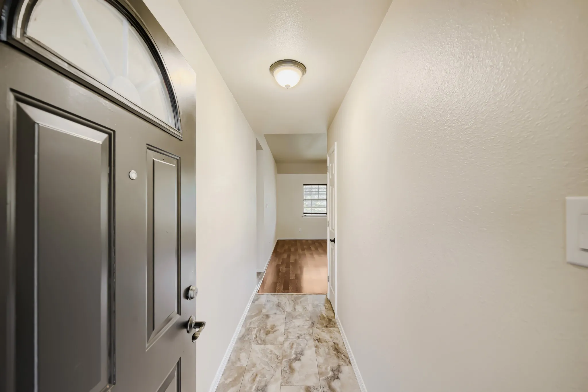 Entryway featuring a dark paneled door with an arched transom window, flush mount lighting, textured light-toned walls, and mixed flooring with stone-look tile transitioning to wood-finish flooring
