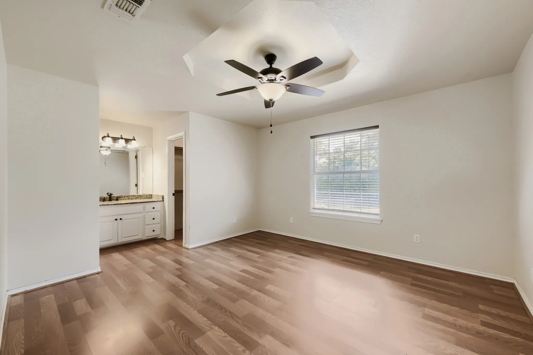 Spacious room featuring wood-finish flooring, a tray ceiling with a ceiling fan, and a window with blinds