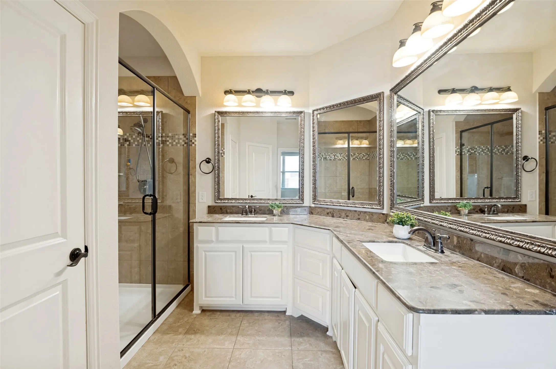 Bathroom featuring a spacious walk-in shower with glass enclosure and decorative tile accent, dual vanities with white cabinetry, integrated sinks, and stone countertops, and multiple framed mirrors with overhead lighting