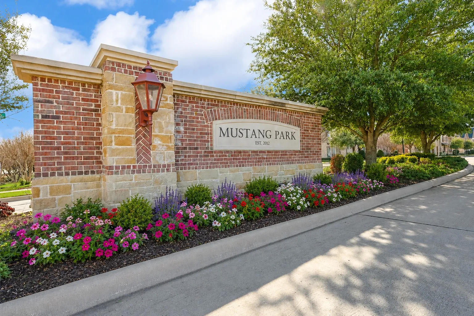 Stone and brick monument sign with a decorative lantern, surrounded by colorful flowerbeds and mature trees