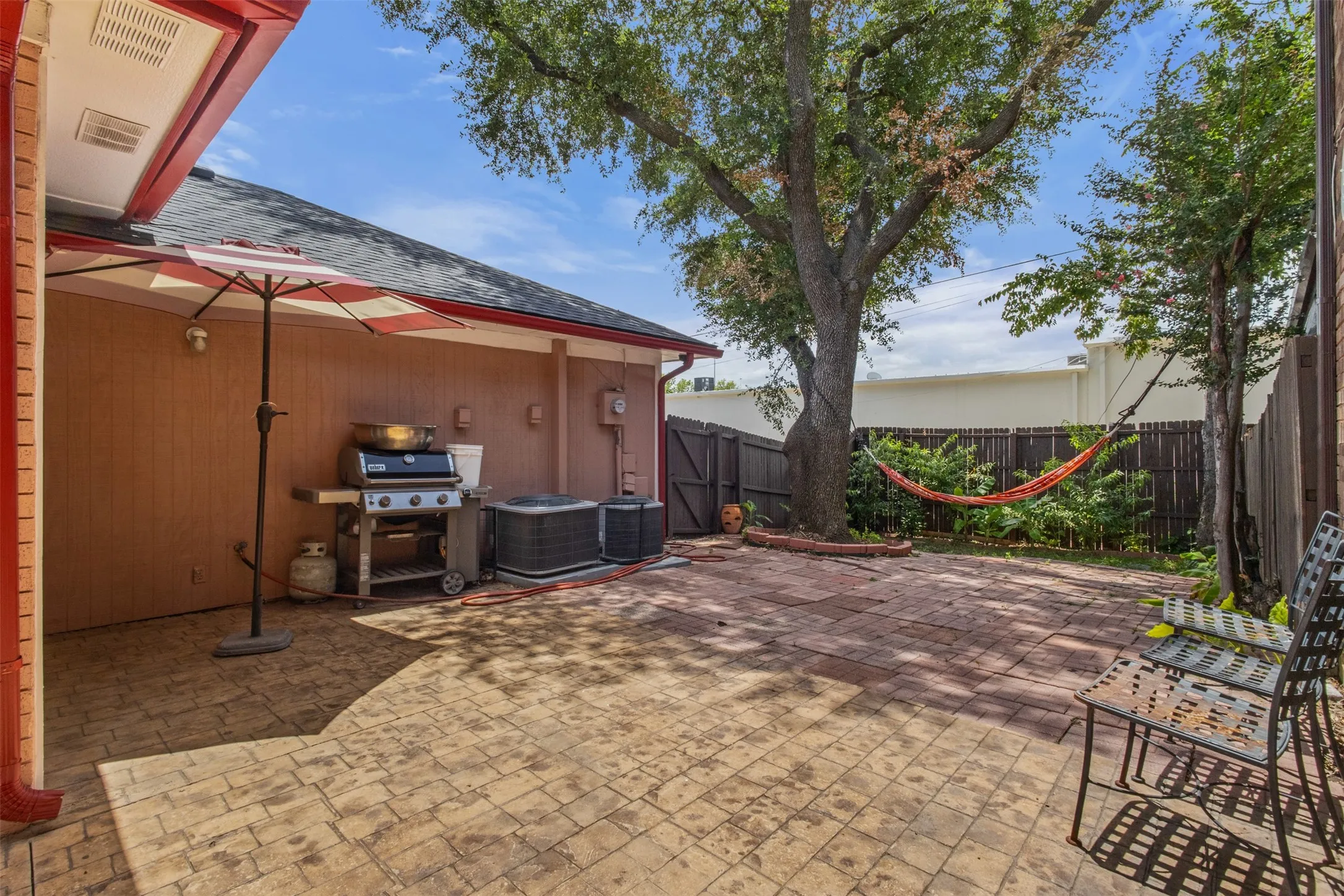 Expansive brick paver patio featuring mature trees, a wooden privacy fence, and a red-trimmed roofline
