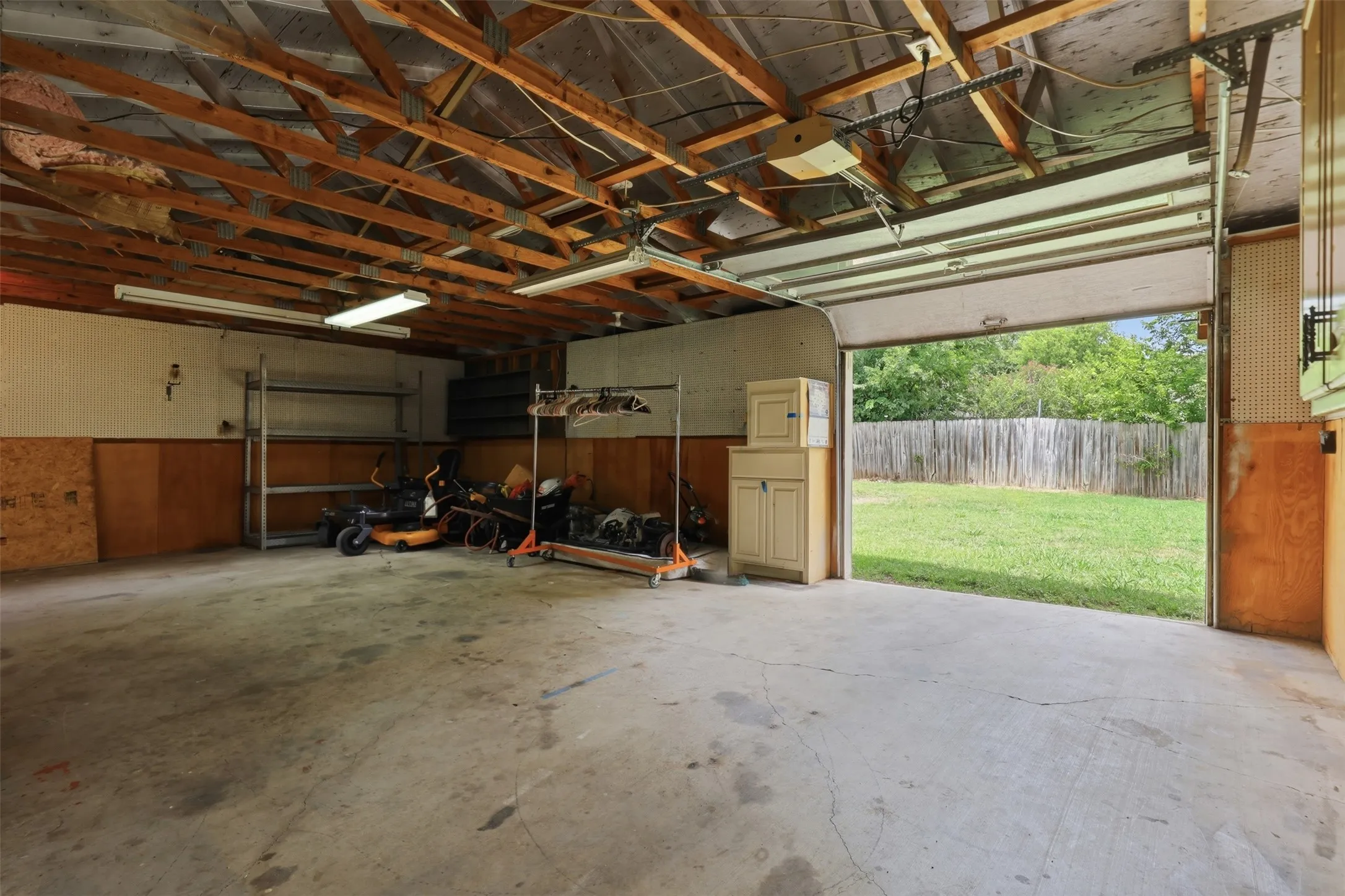 Spacious garage with exposed wood rafters, concrete flooring, fluorescent lighting, wood paneling, and an open garage door leading to a fenced backyard with a manicured lawn