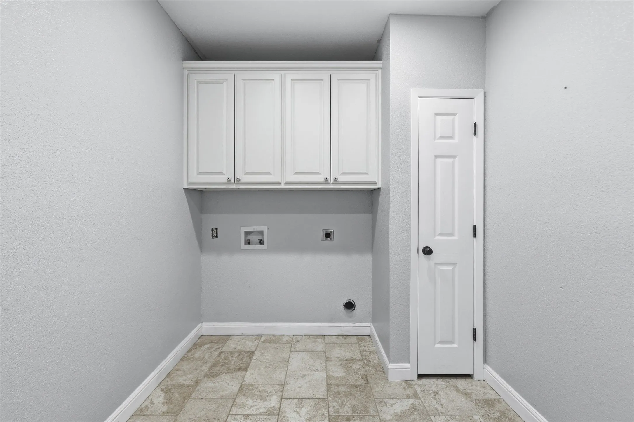 Laundry area featuring light gray walls, built-in white cabinetry, textured tile flooring, and white baseboards