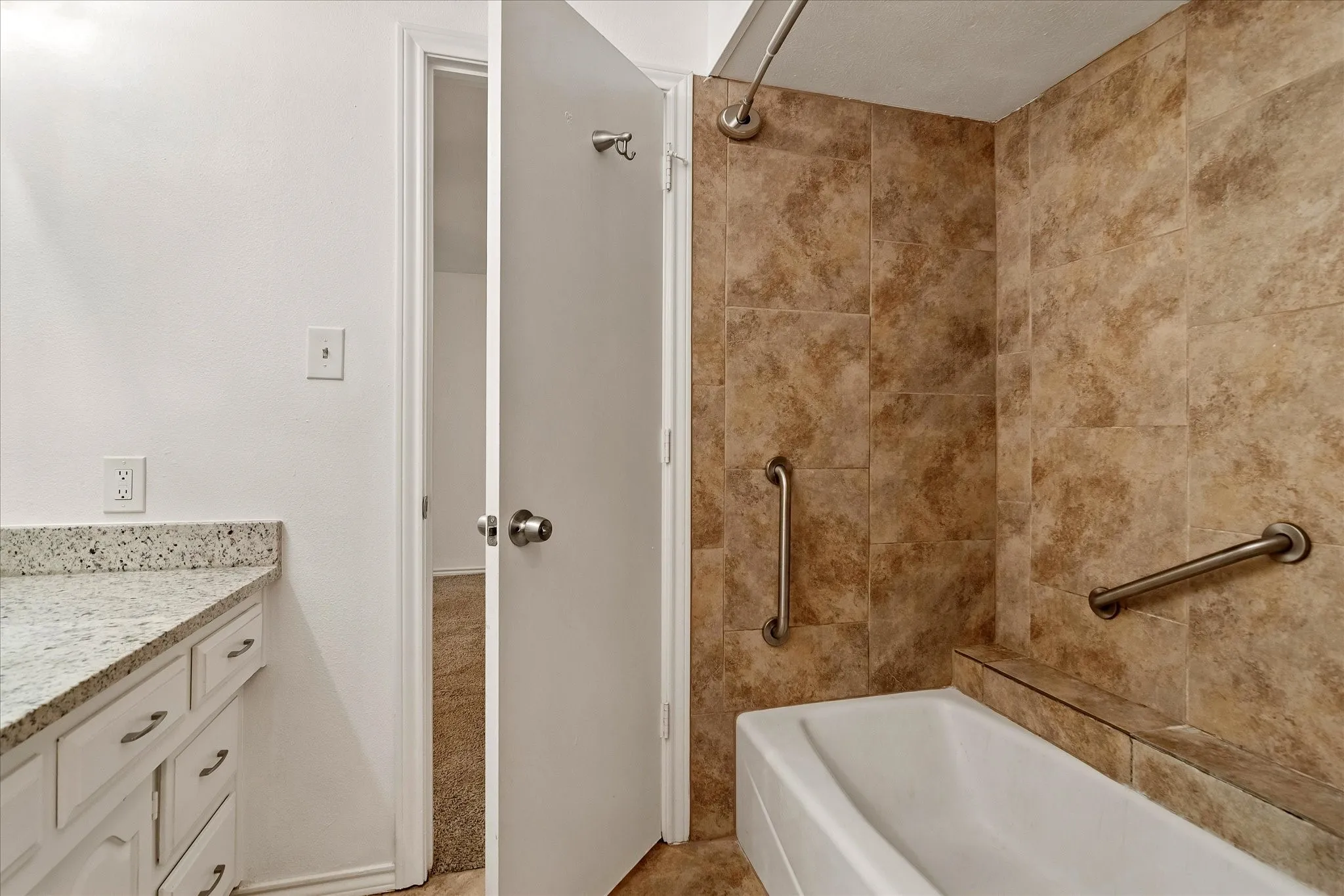 Bathroom featuring a white bathtub with a shower, brown tile surround, two grab bars, and a white vanity with a light-colored speckled countertop