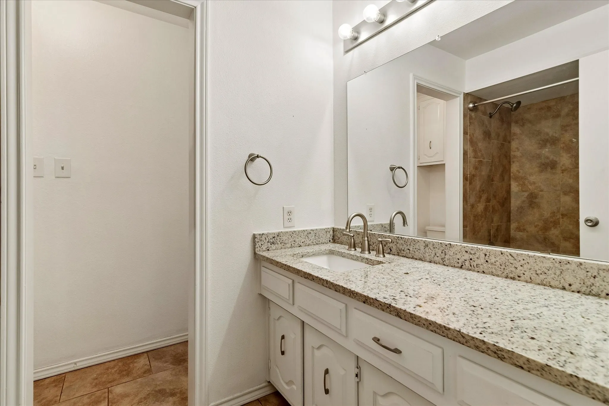 Bathroom featuring a granite-finish countertop, dual-handle faucet, white cabinetry with silver-tone hardware, and a mirrored vanity