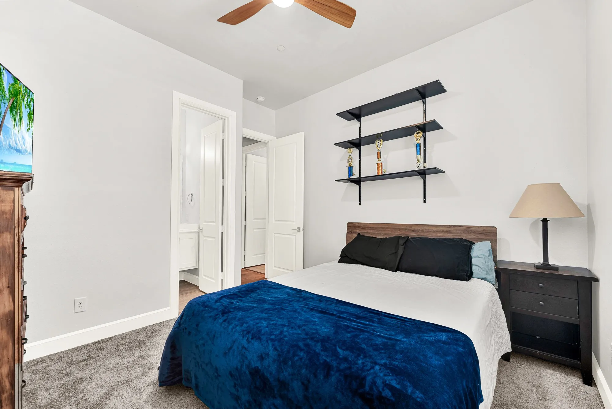 Carpeted bedroom featuring a wooden ceiling fan, white baseboards and closet with a full bathroom next to it.