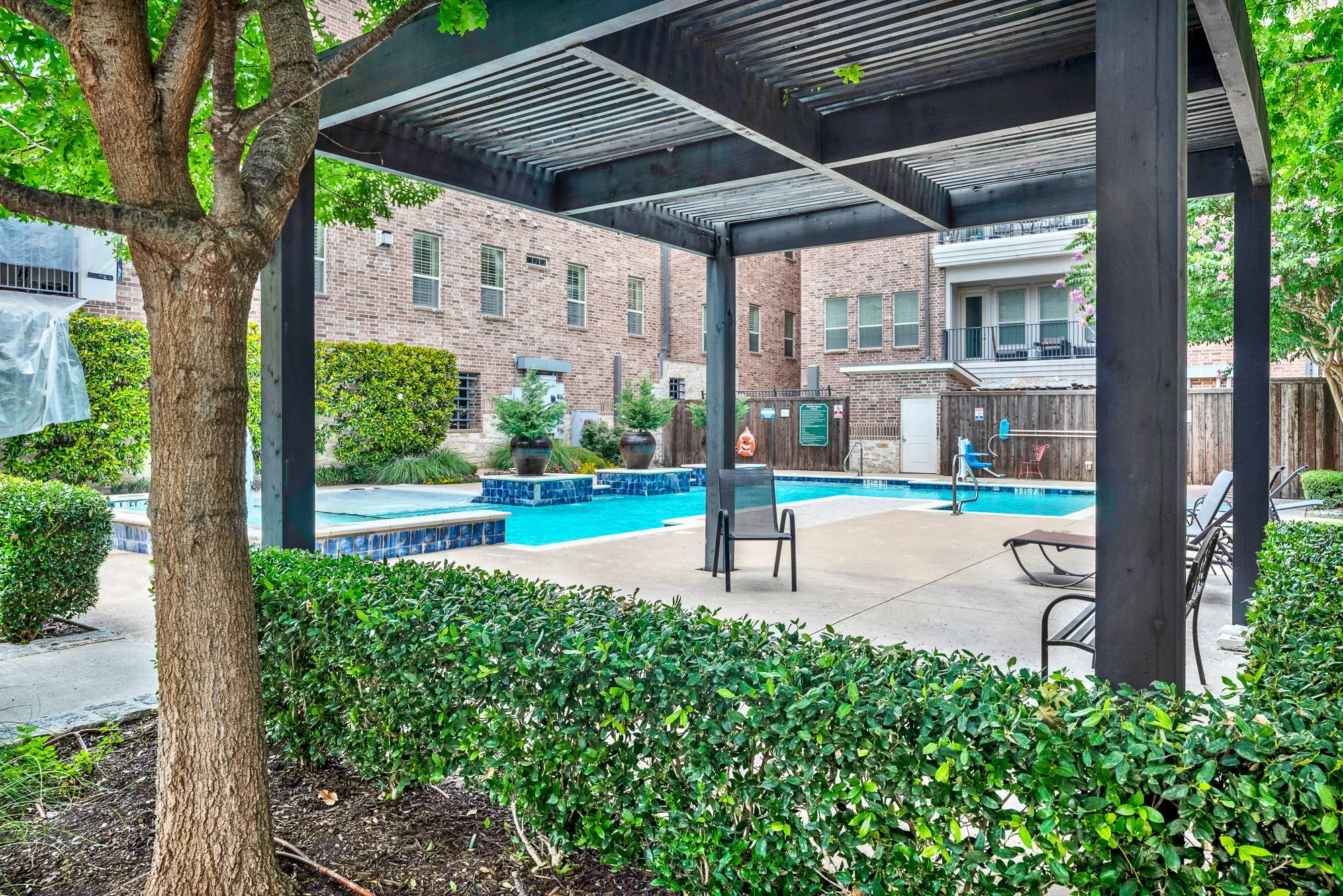 Community pool area featuring a dark-stained wood pergola, rectangular swimming pool, integrated spa, surrounding concrete patio, and manicured hedges