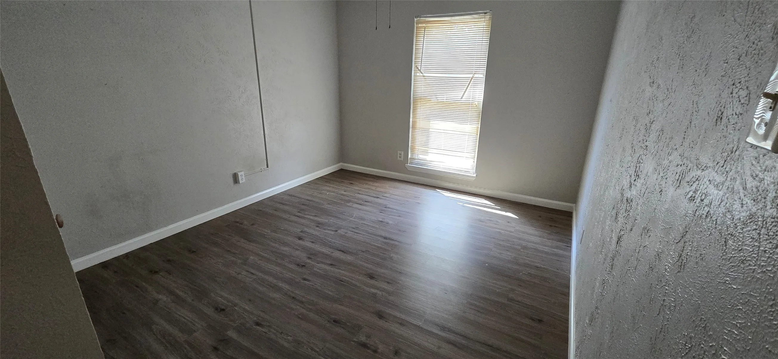 Room with wood-finish flooring, textured walls, and a window with blinds
