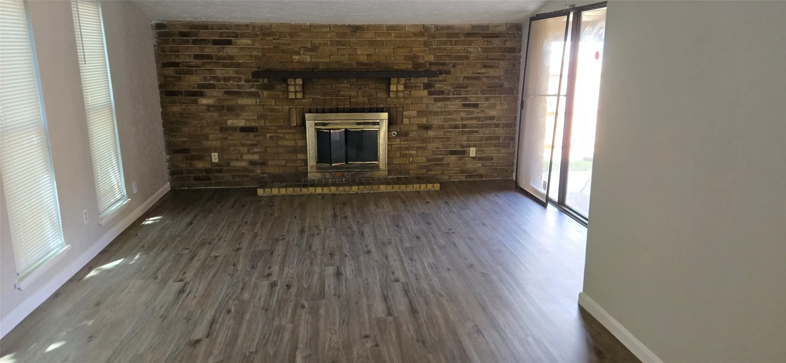 Spacious room featuring wood-finish flooring, a brick accent wall with a fireplace and mantel, multiple windows with blinds, and sliding glass doors