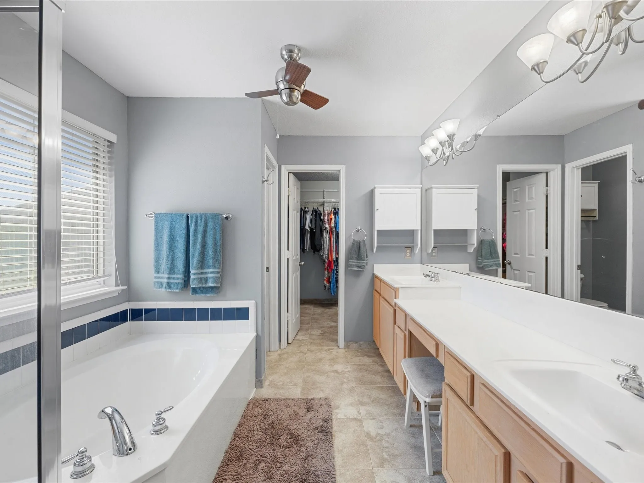 Bathroom featuring a built-in soaking tub with blue accent tile, a double vanity with white countertops, and a ceiling fan