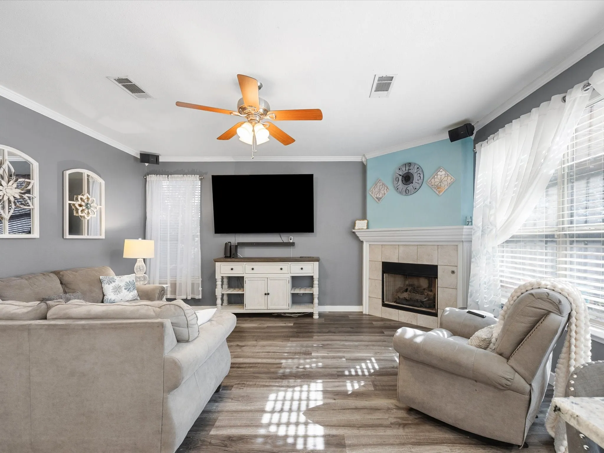 Living area featuring wood-finish flooring, a corner fireplace with a tile surround and white mantel, a ceiling fan, and crown molding