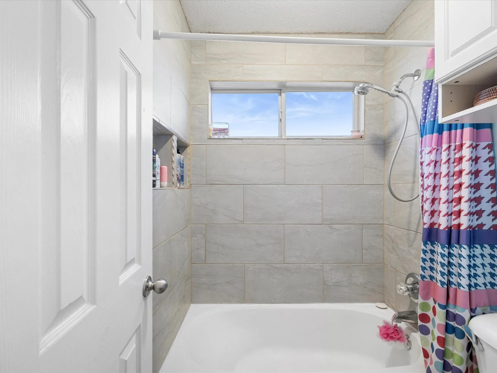 Bathtub with light-toned tiled surround, featuring a built-in shower niche, a horizontal window, and a shower fixture with a handheld wand